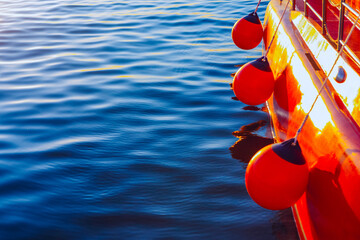 Vibrant red buoys dangle from railing of sleek red boat on calm blue waters, black tops contrasting against glossy hull. Sunlight casts shimmering reflections on gentle ripples, evoking nautical scene