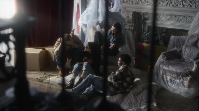 Authentic sneak shot group of young people sitting on mattresses and hanging out in secret hideout together at abandoned house with furniture in foreground, copy space