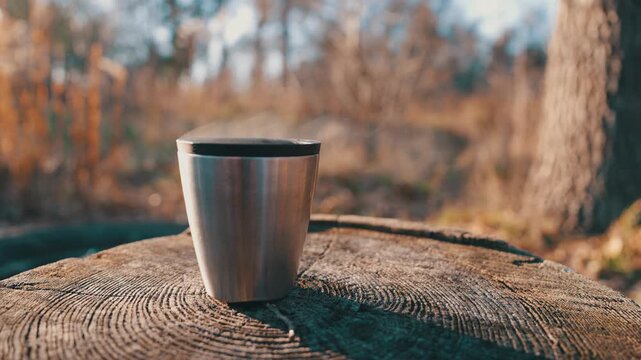 Stainless Steel Thermos Cup Steaming on Tree Stump in Autumn Forest | Hot Drink During Camping Trip at Sunset | Outdoor Adventure and Wilderness Recreation Concept