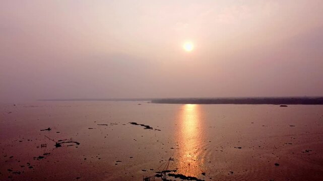 High aerial view of a vibrant, hazy sunset reflecting a golden path across a wide, shallow, and reddish body of water with scattered debris, creating a peaceful, warm atmosphere.