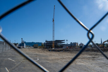 A tall yellow crane rises above a partially constructed steel-frame building, viewed through the diamond pattern of a chain-link fence. The ground is layered with dirt and gravel, scattered 