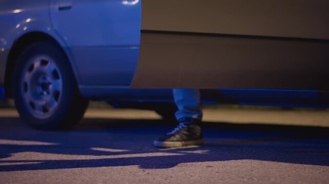 night curb closeup underbody foot near open door, adult rideshare driver in sneakers and jeans pausing beside silver sedan, blue neon and streetlight glow, lowangle shot revealing shadowed panel