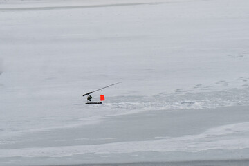 A winter ice fishing setup featuring a fishing rod mounted on a small platform with a bright orange flag, marking a tip-up mechanism positioned over a drilled hole in the ice. Footprints i