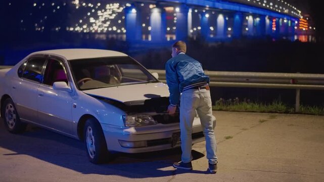 white commuter fixing car under bridge, late night roadside repair scene with neonblue bridge lights, open hood on silver sedan, man in blue jacket using wrench and flashlight, focused expression