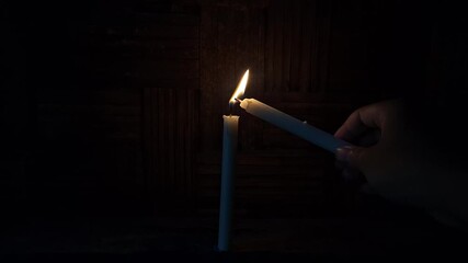 A hand holding a lit candle transfers its flame to a freestanding white candle against a dark, woven bamboo backdrop. Symbolizing shared hope, tradition, and guidance.