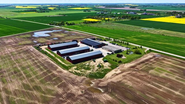 Aerial view of a large farm facility surrounded by wide agricultural fields, with storage barns, access roads, green crop areas, and patches of soil after recent rain, captured in bright daytime.