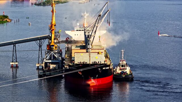 Cargo ship being loaded at an industrial port with cranes and a tugboat assisting alongside, with smoke rising from the vessel and calm blue water surrounding the scene.