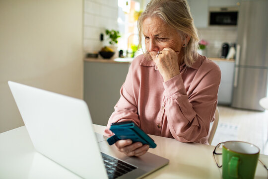 Senior woman looking concerned at phone and laptop at home
