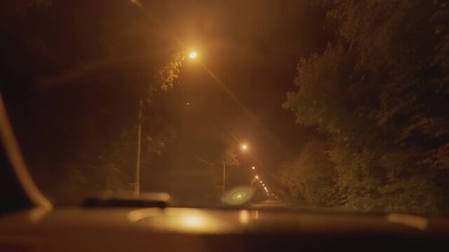 night street lamp glow through mist, warm amber illumination on quiet suburban avenue, windshield dotted with droplets, interior dashboard silhouette hints at latenight rideshare shift, trees
