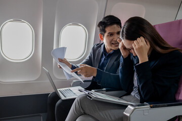 Two Asian business professionals in a heated discussion during a flight. man points at documents with frustration, while the woman covers her ears in stress, sitting in airplane seats with a laptop.