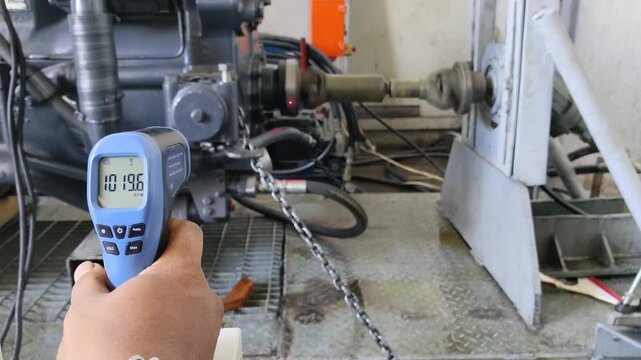 Digital laser tachometer measuring the RPM of a rotating industrial drive shaft, showing a reading of 1021.9 on a blue handheld device in a machinery workshop environment.