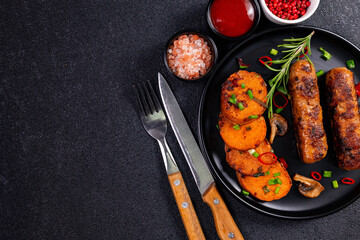 Grilled sausage served with roasted potatoes and spices on a black plate, with utensils and condiments placed nearby on a dark surface