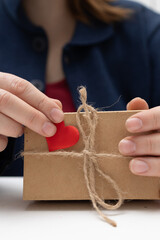 Hands decorate a simple brown gift box. A small red heart is added to the box. The box is also tied with natural brown string. Presented for Valentine's Day on a white surface.