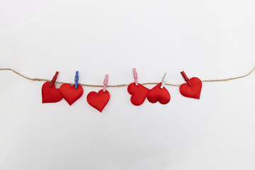 Several red heart shapes are attached to a rustic string with different colored clothespins against a plain white background. This is meant as digital content for Valentine's Day.