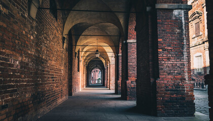 Covered brick archway corridor with hanging lamps and columns, leading to a street.