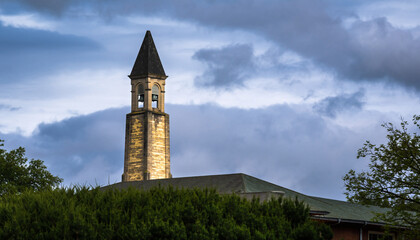 A tall church steeple rises above green trees under a cloudy sky.