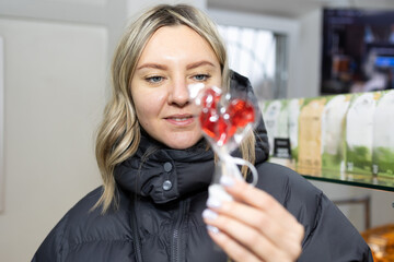 A blonde woman in a black puffer jacket holds and looks at a red, heart-shaped lollipop wrapped in plastic. She is inside a store during the day.