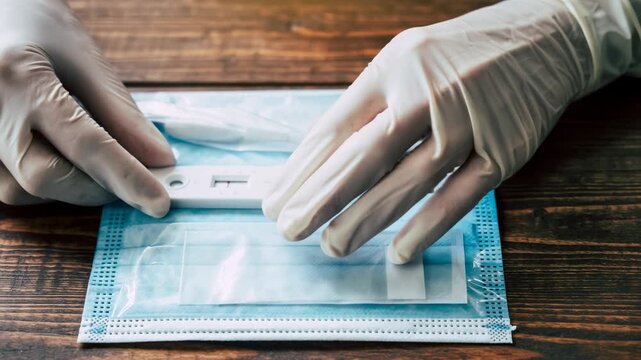 Gloved hands holding rapid test kit on face mask, highlighting pandemic preparedness, healthcare safety, medical glove use, hand protection, and test kit application on wooden table surface