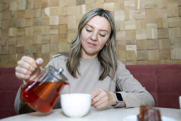 A young woman with blonde hair pours red tea from a clear teapot into a white cup. She sits at a table in a cafe with a wood block wall and a dark red banquette.