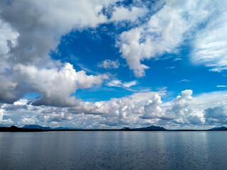 A wide, serene lake reflects the dynamic sky above, with distant mountains creating a subtle horizon line under a dramatic cloudscape