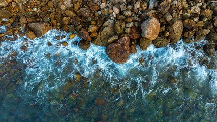 Angle drone shot capturing the beautiful contrast between the deep blue sea foam and the brown textured boulders along the shoreline, creating a natural abstract pattern.concept nature