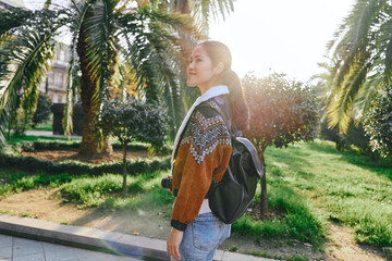 Fototapeta premium Woman with backpack walking along a sunny park path, casual outfit, palm trees in the background, natural light, candid outdoor moment, travel vibe, serene greenery