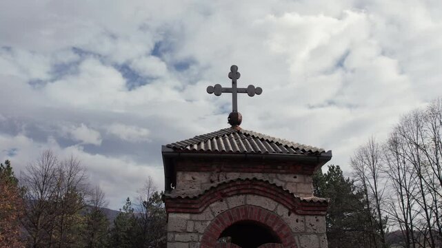 Close-up shot of the Orthodox cross atop the Church of St. John the Baptist in Turbe, Bosnia and Herzegovina, with detailed brickwork and a dramatic cloudy sky above the surrounding autumn trees.