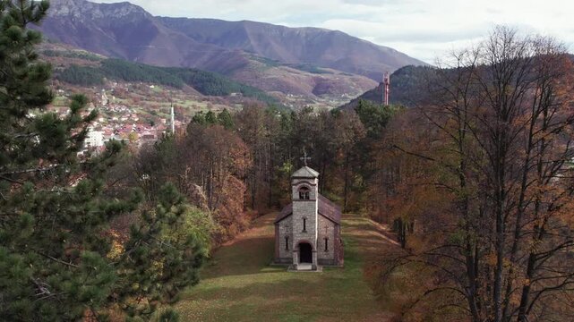 Aerial view of a small Serbian Orthodox stone church surrounded by autumn forest near Turbe, Bosnia and Herzegovina, with mountains and scattered village houses in the background.