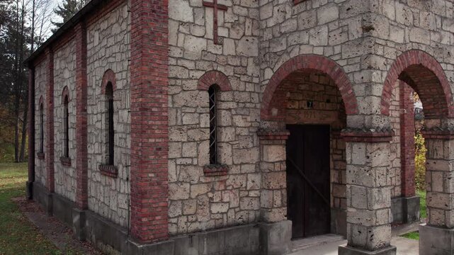 Detailed view of the stone and red-brick entrance of an Orthodox church in Turbe. The historic architecture, arched doorway and window frames stand against a soft autumn forest background.
