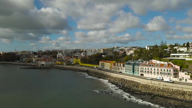 Aerial view of Caxias and Paco de Arcos coastline showing shoreline, coastal road traffic, buildings and calm Tagus river water under a cloudy sky.