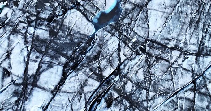Abstract overhead drone shot revealing fractured glacier textures, dark mineral lines, and a small pool of blue meltwater cutting through the icy surface.