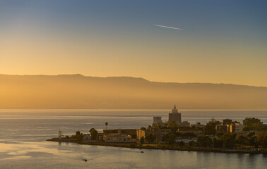 Golden hour lighthouse and coastal fortress on a quiet peninsula above a calm sea with misty mountains and soft haze cinematic seascape travel photography capturing serenity wonder and distant flight