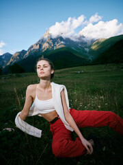 Naklejka premium Outdoor scene of a woman in a white crop top and red pants relaxing on a grassy meadow with rugged mountains and a blue sky, soft sunlight and serene alpine scenery