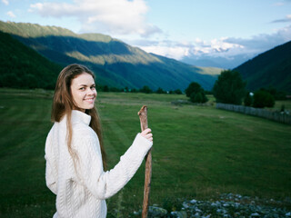 Naklejka premium Woman holding a rustic stick in a green meadow with rolling hills and distant mountains, wearing a warm sweater, bright smile, outdoor scenery and tranquil countryside.