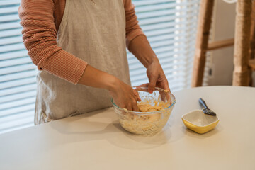 Woman in linen apron mixes dough in a glass bowl on a white table in a bright kitchen