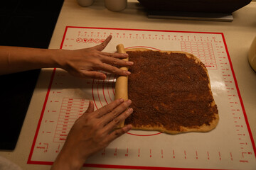 Hands rolling cinnamon-filled dough on a marked pastry mat preparing a sweet treat