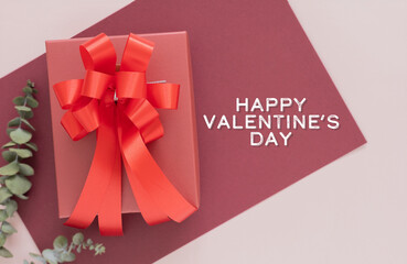 Happy Valentine's Day text next to a red gift box with a large ribbon. A romantic top view flat lay on burgundy and pink backgrounds with eucalyptus for love concepts.