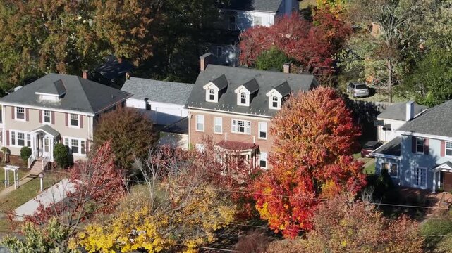 Sweeping drone footage over a picturesque American suburb during peak autumn reveals neat houses, tree-lined streets, and a patchwork of crimson, gold and orange foliage under bright midday light.
