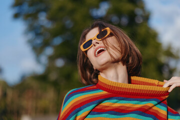 Woman smiling in a colorful striped sweater and yellow sunglasses outdoors, short brown hair, playful pose pulling collar, sunny lifestyle portrait for fashion, travel and summer joy.