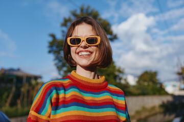 Young woman in a rainbow sweater and yellow sunglasses smiling outdoors under a sunny blue sky....