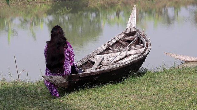 Back view of a peaceful Bangladeshi girl with long black hair, wearing a traditional Salwar Kameez, sitting quietly on an old wooden boat by a calm resort lake. Serene rural scene.