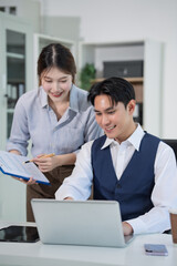 Obraz premium Two Asian business colleagues collaborate in a modern office, reviewing documents and working on a laptop together, showing teamwork, communication, productivity, and professional cooperation