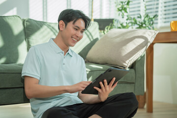 A smiling Asian man relaxes on a sofa using a digital tablet at home, enjoying online content, technology, and comfortable lifestyle moments in a modern living room environment