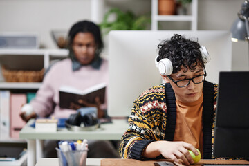 Young adult man with curly hair and glasses wearing headphones working at computer while holding...