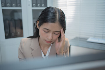 Fototapeta premium Young Asian businesswoman sitting at an office desk, holding her head in frustration while facing a computer, expressing work stress, pressure, burnout, career challenges, emotional straincorporate