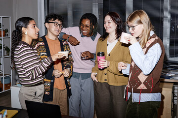 Group of diverse young adults standing together laughing and drinking coffee in office setting, multiethnic friends socializing and enjoying casual conversation during break