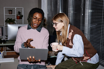Young adult Black man showing laptop screen to young adult Caucasian woman holding mug, both...