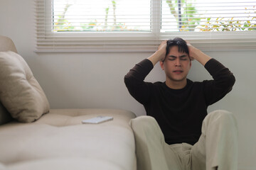Obraz premium Stressed young man sitting on floor with hands on head, experiencing pain and emotional distress, feeling overwhelmed
