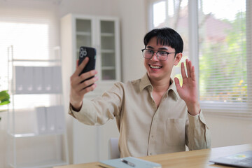 Happy young man holding mobile phone, smiling and waving during an online video conference in a home office