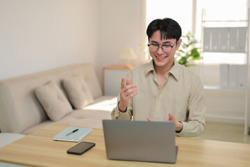 Asian man working from home, smiling and gesturing during a video call on a laptop, engaging in remote work
